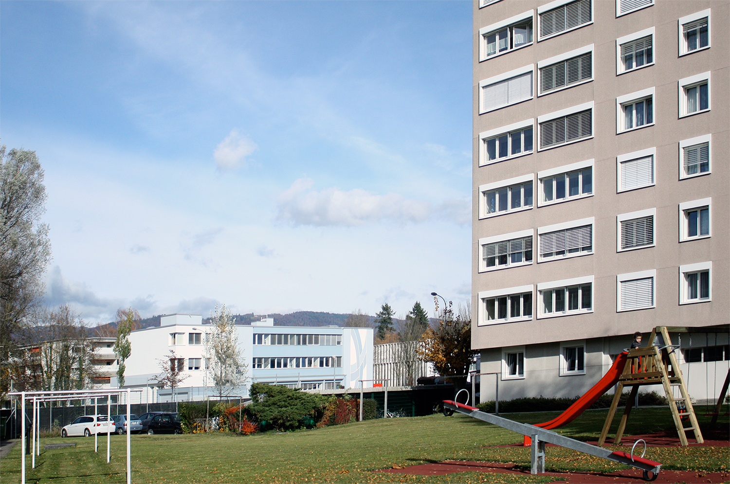 Mehrstöckiges Wohnhaus mit Spielplatz und Jurakette im Hintergrund.
