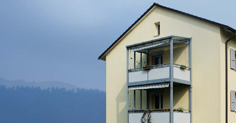 Cremefarbenes Wohnhaus mit Balkonen der Siedlung Bischofkänel vor Hügelkulisse.