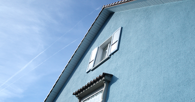 Wohnhaus der Siedlung Mettfeld mit hellblauer Fassade und weissen Fensterläden vor blauem Himmel.